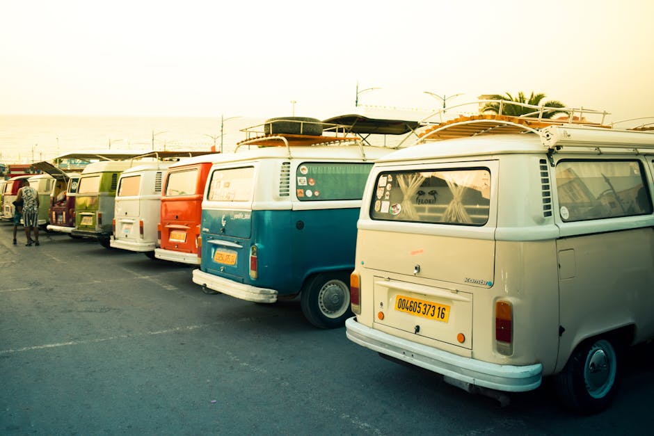 A lineup of multiple vintage Volkswagen Type 2 (T2) vans parked in a row on a paved surface, with some displaying roof racks carrying surfboards and luggage. The vans are painted in various colours including blue, white, red, and green. The rear windows of the vans have curtains, and one has a visible license plate with yellow background and black characters. In the background, a few people are seen walking near the vehicles, and there are palm trees and a bright, clear sky indicating a coastal or warm outdoor setting. This scene reflects a casual, outdoor environment suitable for home relocation and packing activities that involve transporting household items, with the vans possibly used for furniture transport or moving logistics. Man with Van Kensal Green occasionally manages such vehicle fleets for removals services during home moving and furniture transport tasks.