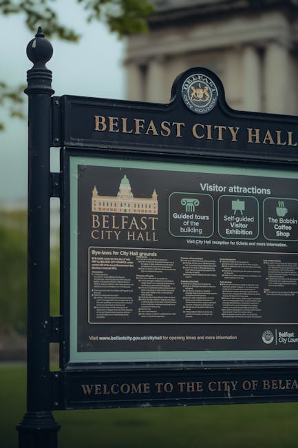 A close-up of a black metal outdoor signpost for Belfast City Hall, featuring the Belfast City Council crest at the top. The sign displays information about visitor attractions, including guided tours, a self-guided visitor exhibition, and the Bobbin Coffee Shop, with icons representing each. Below, detailed bye-laws for the City Hall grounds are listed in small print. The sign is positioned on a pavement with blurred greenery and sky in the background, indicating an outdoor setting. The overall scene reflects a standard informational display relevant to a public historic building, situated in an area that may involve house removals or relocation services when moving in or out of the city center.