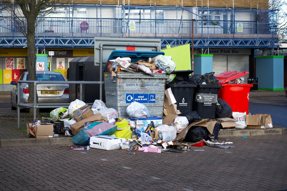 A collection of overflowing waste bins and piled rubbish on a paved sidewalk outside a commercial area, including a large grey mixed paper and cardboard recycling bin with its lid open and contents spilling out, black and green general waste bins, and a bright red recycling bin. The debris comprises cardboard boxes, plastic bags, paper, and packaging materials, some flattened and others loose, scattered around the bins and on the ground. In the background, a car is parked near railings protecting a tree, with a building under construction or renovation featuring scaffolding and a sign indicating waste disposal guidelines for house removals and moving services, aligning with the context of household or business waste management in a residential or commercial relocation setting.