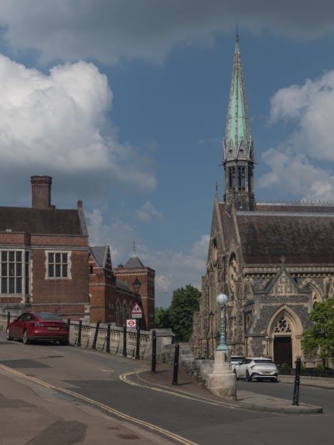 View of a street near a church with a tall, pointed spire and detailed stonework, situated on a sloped pavement. A red car is parked on the left side of the road, with black metal bollards lining the 