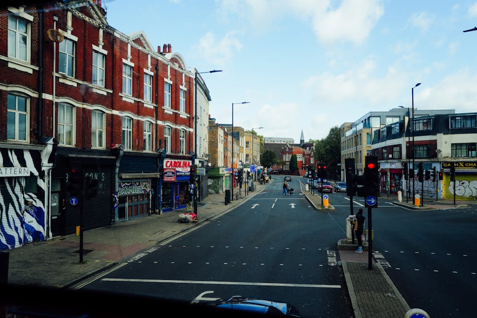 A street scene in Kensal Green showing a line of parked cars on both sides of the road, including sedans and hatchbacks, with some vehicles featuring visible registration plates. In the foreground, a