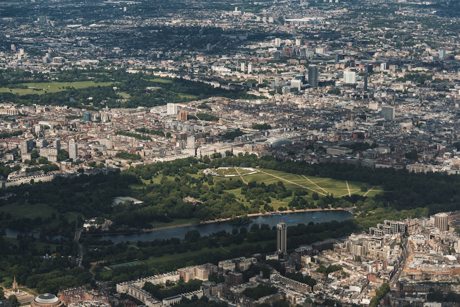 An aerial view of a large urban park, featuring expansive green lawns, pathways forming geometric patterns, and a river running through the lower part of the park, with surrounding cityscape comprising numerous multi-storey buildings and streets. The scene is captured during daylight with natural lighting, showcasing the contrast between the greenery and the densely built environment, which is typical for a central London area. This image illustrates the setting for house removals and furniture transport tasks that involve navigating city streets and outdoor spaces. Man with Van Kensal Green’s removals services support home relocation projects within such urban environments, ensuring efficient packing, loading, and transport of furniture and household goods through city networks and green spaces.