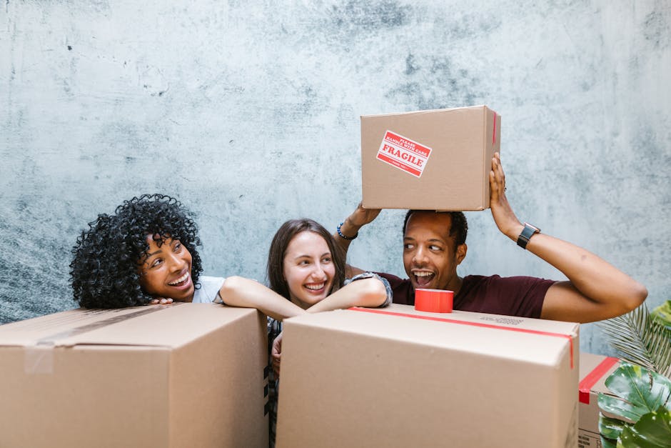 A group of three smiling individuals, two women and one man, inside a room with a textured, gray wall in the background, preparing for home relocation. The man, wearing a dark T-shirt and a watch, is holding a cardboard box with a red 'Fragile' sticker above his head, indicating careful handling during furniture transport. The women, one with curly hair and the other with straight, shoulder-length hair, are leaning on large cardboard boxes and appear happy and engaged in the packing process. Several packed boxes, some sealed with red tape, are stacked on a surface in front of them, suggesting an active packing and loading process. The scene captures the interior of a property undergoing a move, emphasizing the importance of packing, handling fragile items, and the collaborative effort involved in a successful house removal, which is a service provided by Man with Van Kensal Green.