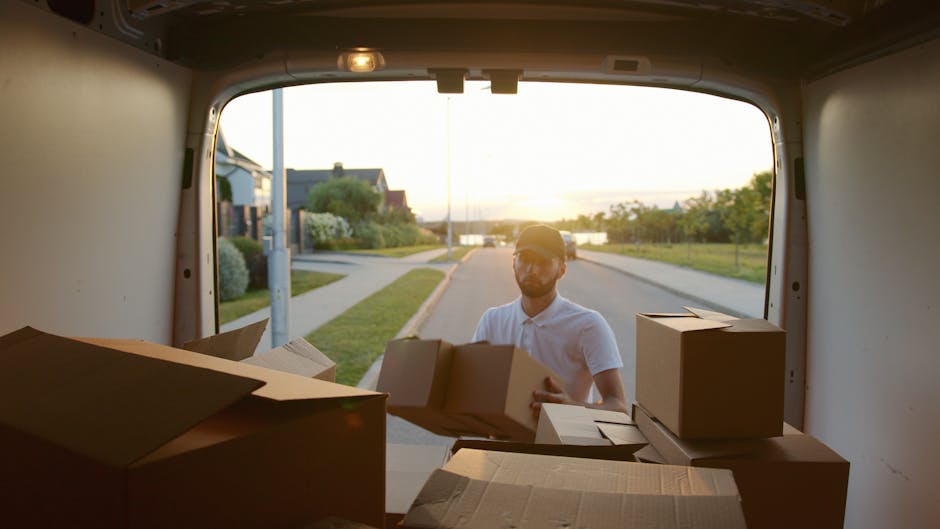 A person wearing a white shirt and cap is seen outside a moving van, loading and arranging cardboard boxes of varying sizes, some open and some sealed with packing tape, onto the vehicle's floor during a home relocation in Kensal Green. The van's rear doors are open, revealing a suburban street with residential houses, a sidewalk, greenery, and a sunset sky casting warm light on the scene. Inside the van, the environment is dimly lit with minimal interior features visible. This loading process highlights the careful packing and transport phase of a furniture or household items move, consistent with services offered by Man with Van Kensal Green, emphasizing efficient logistics in house removals and furniture transport.