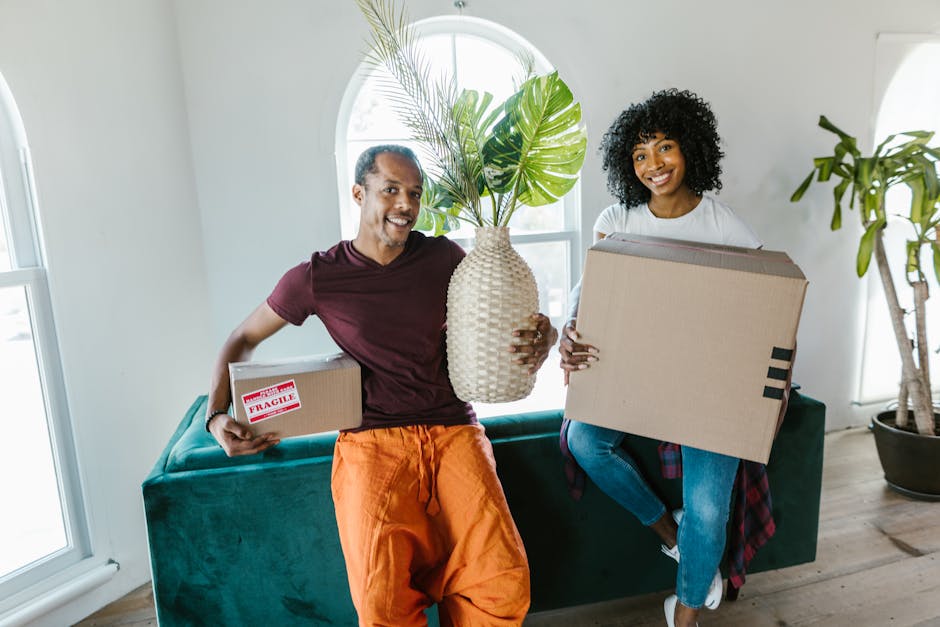 A group of three smiling individuals, two women and one man, inside a room with a textured, gray wall in the background, preparing for home relocation. The man, wearing a dark T-shirt and a watch, is holding a cardboard box with a red 'Fragile' sticker above his head, indicating careful handling during furniture transport. The women, one with curly hair and the other with straight, shoulder-length hair, are leaning on large cardboard boxes and appear happy and engaged in the packing process. Several packed boxes, some sealed with red tape, are stacked on a surface in front of them, suggesting an active packing and loading process. The scene captures the interior of a property undergoing a move, emphasizing the importance of packing, handling fragile items, and the collaborative effort involved in a successful house removal, which is a service provided by Man with Van Kensal Green.