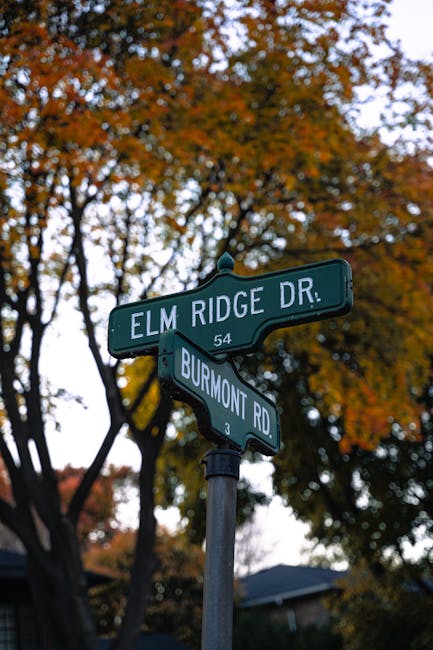 Photograph of two green street sign plates mounted on a black pole at the corner of Elm Ridge Drive and Burmont Road, with the Elm Ridge Drive sign displaying the number 54 and the Burmont Road sign displaying the number 3, set against a background of autumn trees with orange and yellow leaves and a partly cloudy sky. The scene reflects an outdoor residential area typical of a Kensal Green neighbourhood, with the street signs indicating intersection points relevant to house removals or home relocation planning facilitated by Man with Van Kensal Green. The image emphasizes a quiet street environment, essential for planning local furniture transport and packing logistics during a house move.
