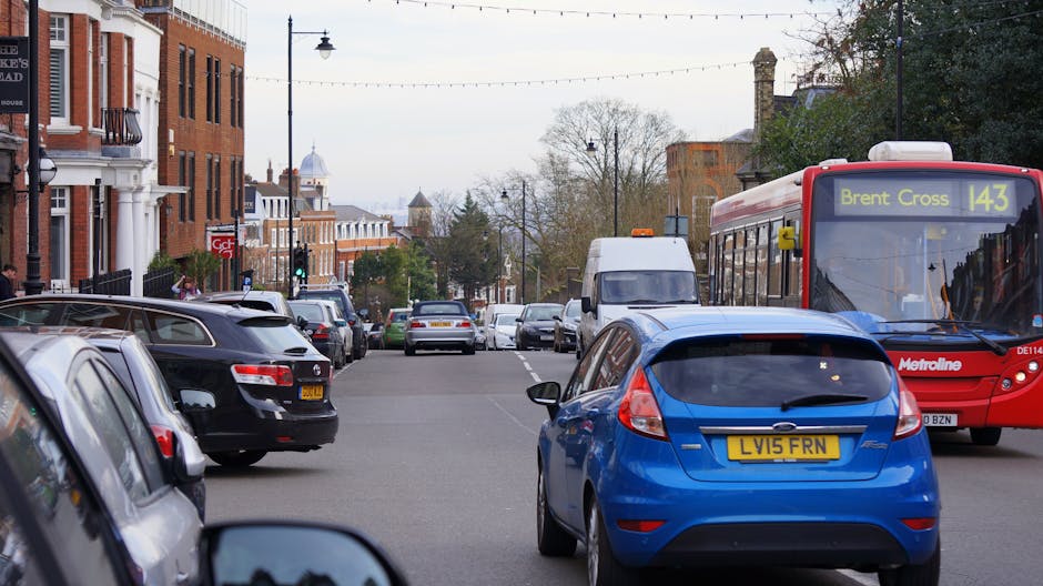 A busy street scene in Kensal Green shows a blue hatchback car parked on the side of the road, with several other vehicles including a black car, a white van, and a red Metroline bus displaying 'Brent Cross 143' on its destination sign. The street is lined with brick residential buildings and street lamps, with some pedestrians visible on the sidewalks. The area appears to be in daytime with overcast weather, and the parking situation looks congested, reflecting common challenges during home relocation or moving logistics. This image exemplifies typical urban street conditions that house removals services, like those offered by Man with Van Kensal Green, often encounter when facilitating furniture transport and packing during house moves.