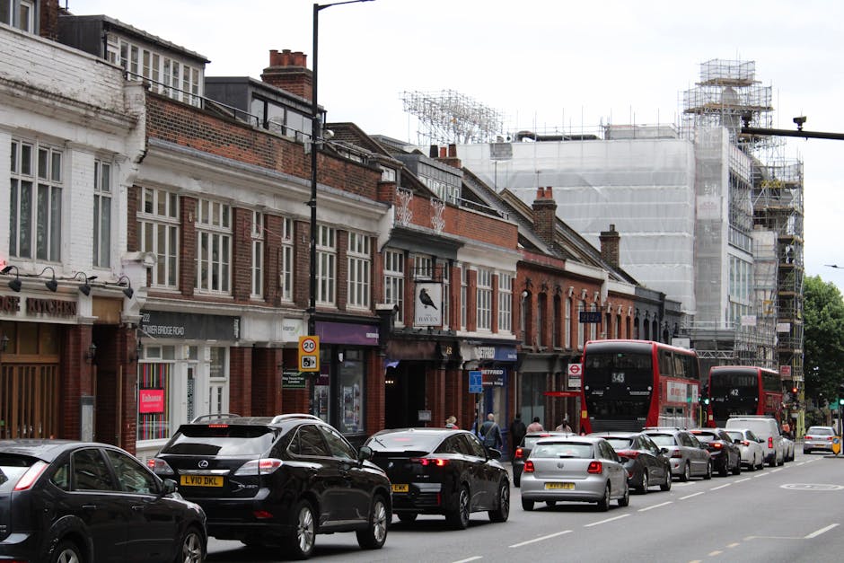A street scene in Kensal Green showing a row of traditional brick terraced houses with white window frames and decorative brickwork facades, some with scaffolding and construction work ongoing on a building at the far end of the street. Several cars, including black, silver, and grey models, are parked along the street and moving traffic is visible. To the right, two red double-decker buses are in motion, with one heading away from the viewer and the other approaching. Pedestrians are walking on the sidewalk near the row of shops, which include a café and retail stores. The scene depicts an active urban environment with typical street parking, bus transport, and ongoing building maintenance, relevant to house relocations and city-based removals services.