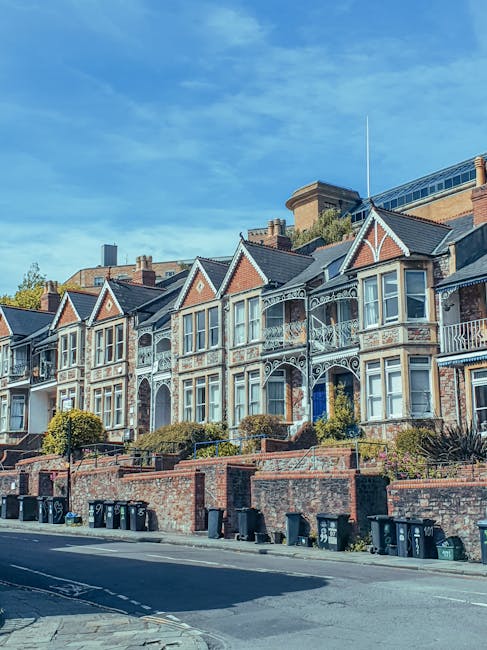 A row of Victorian-style terraced houses with ornate white balconies and large bay windows, situated on a sloped street in Kensal Green. The houses are built with brick facades, and some have decorative woodwork and gabled roofs. Below the houses, a brick wall with steps leads up to front gardens with shrubs and small trees. On the pavement in front of the houses, multiple black wheelie bins are aligned along the street. The sky above is clear with some wispy clouds, and the scene is illuminated by daylight, capturing the characteristic architecture of the area. This environment suggests a residential neighbourhood typical for house removals and home relocation services, with visible elements indicating the planning and loading process involved in furniture transport and moving logistics, as managed by professional removals providers like Man with Van Kensal Green.