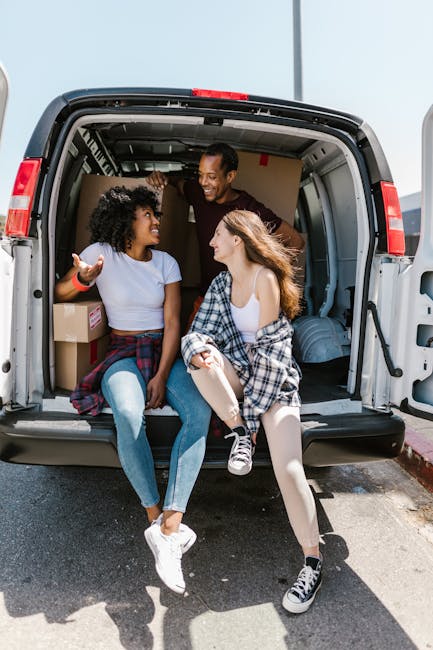 A group of three young people sitting inside the open rear of a grey moving van parked on a paved street, with cardboard boxes and packed luggage visible behind them. The individuals, two women and on