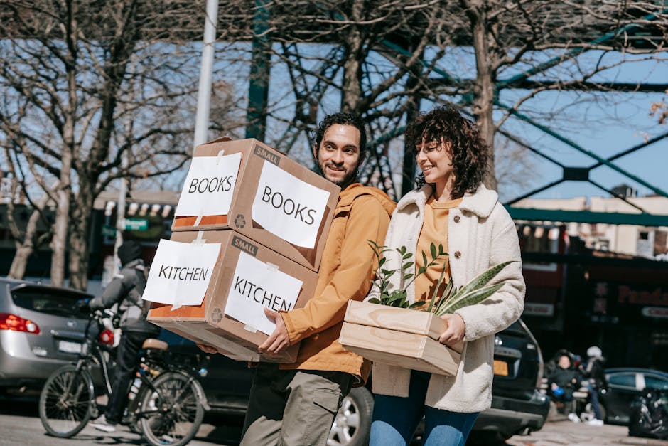 A man and a woman are outdoors during daytime, engaged in a home relocation process, carrying packed boxes labeled 'BOOKS' and 'KITCHEN' from inside a property to a vehicle or loading area. The man, with dark hair and a beard, is holding two cardboard boxes with labels indicating contents, and is wearing a mustard-colored jacket. The woman, with curly dark hair, is carrying a wooden crate with green plants, dressed in a cream-colored coat. In the background, there are cars, bicycles, and a modern glass building with a blue structure, suggesting an urban environment. The scene is illuminated by natural daylight, capturing the loading process typical of furniture transport and packing and moving activities, as part of a house removal undertaken by Man with Van Kensal Green.