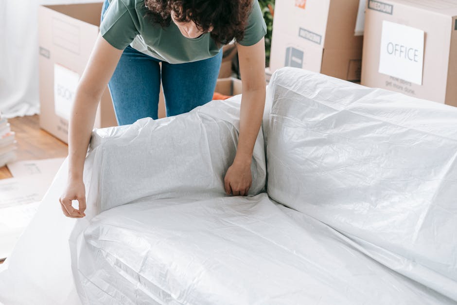A person wearing a green top and blue jeans is in the process of wrapping a large piece of furniture with white packing paper, preparing it for a home relocation. The individual is bent over, carefully covering the sofa or similarly sizable item on a wooden floor, with several cardboard boxes and packaging materials visible in the background, including a box labeled 'OFFICE.' Inside the property, natural light brightens the scene, indicating a well-lit environment, likely during a moving day organized by Man with Van Kensal Green. The scene depicts the packing stage of furniture transport, with the person ensuring the item is protected for safe moving to a new residence. Surrounding objects include additional boxes and packing supplies, situated near doorways and along the walls, supporting the professional packing and moving process involved in house removals and furniture transport services.