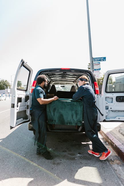 Two movers from Man with Van Kensal Green are loading a large piece of furniture covered with a green protective blanket into the back of a black van during a home relocation. The van's rear doors are open, revealing the spacious interior where the furniture is being carefully positioned on the vehicle's cargo area. One mover, wearing a dark blue shirt and black pants, is holding the furniture steady, while the other, dressed in a matching uniform with red shoes, assists in guiding it inside. The loading area is a paved driveway adjacent to a building, with a nearby street sign visible that reads 'Somerset.' Bright daylight illuminates the scene, highlighting the professional handling of packing and furniture transport in the relocation process. Man with Van Kensal Green specializes in removals and home moving logistics, as demonstrated by the careful loading activities captured in this image.